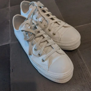 Vita Converse All Star sneakers - Klassiska vita Converse All Star sneakers i lågt snitt med snörning och guldiga öljetter. Skorna har rund tå, platt sula och är tillverkade i skinn med textilfoder. Perfekta för en clean och stilren look.Andvända 2 gånger