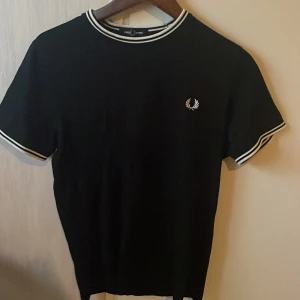 Svart Fred Perry t-shirt med vita detaljer - Svart t-shirt från Fred Perry med klassisk vit logga på bröstet. T-shirten har vita ränder på krage och ärmslut, rund hals och korta ärmar. Perfekt basic med snygga kontrastdetaljer och sportig känsla.