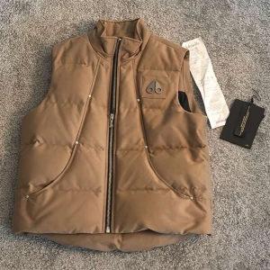 Brun dunväst från Moose Knuckles - Säljer en brun dunväst från Moose Knuckles med coolt broderi på insidan och metallogo på bröstet. Västen har hög krage, två stora fickor framtill och dragkedja. Perfekt för lager-på-lager. Nypris runt 4000kr. Strl XL men sitter mer som L/M. Skick 9,5/10 svinbra skick. Hör av er för mer information osv! 