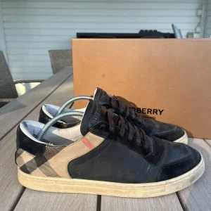 Burberry sneakers med rutigt mönster - Snygga Burberry sneakers i svart skinn och mocka med klassiskt beige och svart rutigt tyg på sidorna. Skorna har svarta snören, vit sula och Burberry-logga på plösen. Medföljer extra vita skosnören och originalkartong. Köpt på cettire för 5000 har kvitto osv 