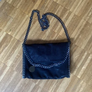 Svart axelväska med kedjedetaljer - Snygg svart axelväska med glansig finish och coola kedjedetaljer runt kanterna och på axelremmen. Väskan har ett lock med magnetknapp och insidan är fodrad med leopardmönster. Perfekt storlek för det viktigaste och ger en edgy vibe till din outfit.