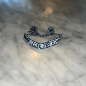 Märkesarmband blått - Snyggt märkesarmband texten i vitt och blå. Armbandet har justerbar knytning med tofsar i ändarna och en cool, casual vibe. Perfekt accessoar för att lyfta din outfit med en touch av lyx.