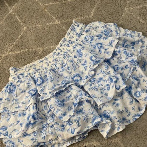 Blommig volangkjol från Hollister - Supersöt kort kjol från Hollister med blått blommönster på vit botten. Kjolen har flera volanger och bred resår i midjan för en bekväm passform. Perfekt för varma dagar och ger en fräsch och somrig vibe.