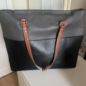 Stor svart och grå axelväska med bruna band - Snygg och rymlig axelväska i svart och grått med bruna axelband. Väskan har dragkedja upptill och flera innerfack för att hålla ordning på dina grejer. Perfekt för dig som vill ha plats för både laptop och annat viktigt.