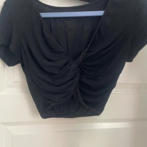 Svart croppad topp med twistad front - Svart croppad topp med korta ärmar och snygg twistad detalj framtill. Toppen har v-ringning och är gjord i ett ribbat material