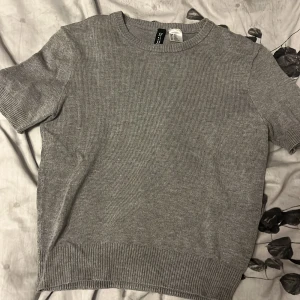 Grå stickad kortärmad tröja Divided - En basic grå stickad tröja från Divided by H&M. Tröjan har korta ärmar, rund halsringning och ribbade muddar vid ärmslut och nederkant. Perfekt för lager-på-lager och funkar året runt. Enkel och stilren design som passar till allt.