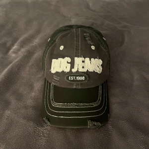 Grön keps från BDG Jeans - Grön keps från BDG Jeans med vit broderad logga och texten 'EST. 1998' framtill. Kepsen har slitna detaljer och kontrastsömmar som ger en edgy streetkänsla. Perfekt accessoar för att lyfta din outfit, även justerbar!😍