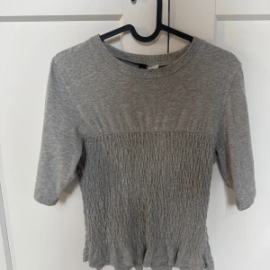 Grå smockad topp från Divided XS - Grå topp från Divided i storlek XS med smockad midja och volangkant nedtill. Toppen har rund halsringning och halvlånga ärmar. Mjuk och stretchig bomullsblandning som sitter snyggt och bekvämt.