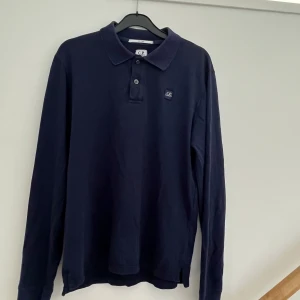 Mörkblå långärmad pikétröja CP Company - Snygg mörkblå långärmad pikétröja från CP Company med klassisk krage och knappslå framtill. Tröjan har en liten logotyp på bröstet och är gjord i mjuk bomull. Använd men i bra skick. Bara har av sig vid eventuella funderingar!😉