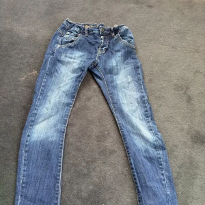 Blå jeansbyxor från Nicky, stl 146 - Snygga blå jeansbyxor från Nicky i storlek 146. Klassisk femficksmodell med ljusa slitningar framtill och bakfickor med dekorativa sömmar. Jeansen har normal passform och är tillverkade i slitstarkt denimtyg.