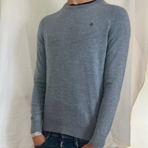 Morris stickad tröja 100% MERINOULL  - Snygg grå tröja från Morris i 100% merinoull. Klassisk rund halsringning och diskret brunt broderat emblem på bröstet. Långärmad och ribbade muddar för en clean look. Perfekt för dig som gillar stilrena och bekväma plagg. Den är ljusblå blandat med lite grå.