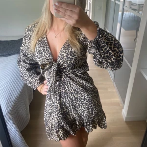 Leopardmönstrad kortklänning med knyt - Snygg kort klänning med leopardmönster och volangkant nertill. Den är aldrig använd endast testad!!! Djup v-ringning och knyt framtill i midjan. Långa puffiga ärmar med resår vid handlederna. 