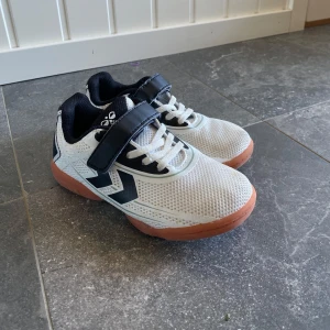 Träningsskor barn 31 - Snygga vita sneakers från Hummel med svart detalj och logga på sidan. Skorna har både snörning och kardborreband för extra bra passform. Ovandelen är i mesh och sidorna har svarta detaljer. Inomhus träningsskor. 