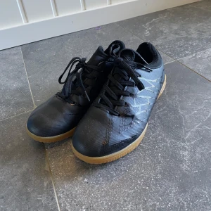 Svarta träningsskor barn 33 - Snygga svarta fotbollsskor med diskret mönster och klassisk snörning. Skorna har en låg profil och en ljusbrun gummisula som ger bra grepp på inomhusplaner. Perfekta för dig som gillar att spela inomhusfotboll. Sparsamt använda. 