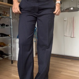 Randiga kostymbyxor från Monki - Mörkblå kostymbyxor från Monki med smala vita ränder. Byxorna har hög midja, raka ben och klassiska hällor för bälte. Snygg dressad look med stängning fram och sidfickor. Perfekta för en stilren och trendig outfit.