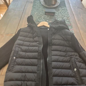 Svart dunväst från Moncler, M - Säljer en svart dunväst från Moncler i storlek M, sprillans ny använd en gång. Västen har quiltad framsida, stickade sidor och rygg samt dragkedja framtill. Klassisk Moncler-logga på ärmen och praktiska fickor med tryckknappar. Perfekt för lager-på-lager och snygg streetstil. 