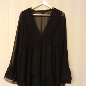 Svart prickig klänning Gina Tricot 40 L - Supersöt svart kortklänning från Gina Tricot med transparenta långärmar, v-ringning och små prickar i tyget. Klänningen har volangdetaljer och spets vid ärmsluten, vilket ger en romantisk vibe. Perfekt för dig som gillar trendiga och feminina plagg.