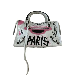 Balenciaga grafitti mini bag 24 cm  - Cool vit handväska från Balenciaga med graffititryck i svart och rosa. Väskan har handtag med flätad detalj, dragkedja framtill och unika handmålade texter som 'PARIS' och 'LEATHER'. Tillverkad i skinn och har svarta metalldetaljer. Perfekt statement piece för dig som vill sticka ut.