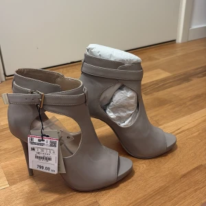 Gråa klackade stövletter med öppen tå - Snygga grå stövletter från Zara i skinn med öppen tå och höga smala klackar. Skorna har coola utskurna detaljer på sidorna och justerbart spänne runt ankeln i silver. Perfekta för dig som vill sticka ut med en modern och stilren look.