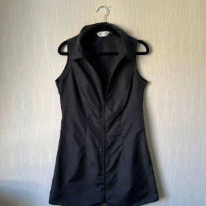 Svart ärmlös klänning från Topshop - Svart kortklänning från Topshop med krage och hel dragkedja framtill. Klänningen är ärmlös och har en lätt figurnära passform. Materialet är slätt och ger en cool, minimalistisk vibe. Perfekt för dig som gillar stilrena och edgy plagg.