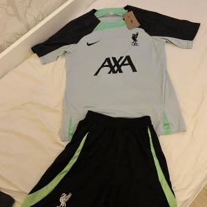 Liverpool Nike fotbollströja ljusblå/svart - Liverpool FC träningsset från Nike med ljusblå kortärmad tröja, svarta ärmar med subtilt mönster och mintgröna detaljer. AXA-logga och LFC-märke på bröstet. Matchande svarta shorts med mintgröna ränder och klubbmärke. Svalt funktionsmaterial.