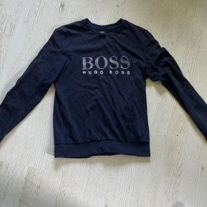 Svart långärmad tröja Hugo Boss - Svart långärmad tröja från Hugo Boss med stort BOSS-tryck framtill i vitt. Tröjan har rund halsringning och är tillverkad i mjuk bomull, perfekt för en clean och stilren look.