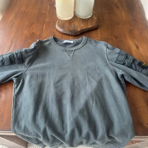 Grå sweatshirt från Stone Island - Säljer en grå sweatshirt från Stone Island med klassisk rund hals och ribbade muddar. Tröjan har unika detaljer på ärmarna och den ikoniska Stone Island-loggan på vänster ärm. Perfekt för dig som gillar streetwear och snygga basplagg.