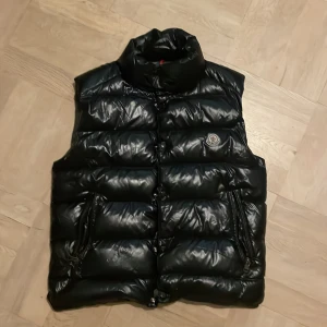 Svart dunväst från Moncler - Säljer en svart, glansig dunväst från Moncler med hög krage och klassisk logga på bröstet. Västen har två dragkedjefickor på sidorna och stängs med både dragkedja och knappar. Perfekt för lager-på-lager och riktigt snygg till streetwear.