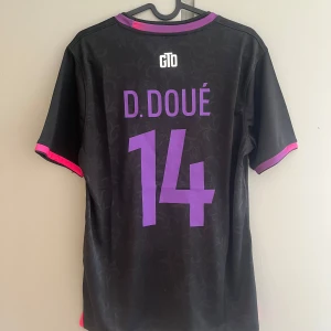 PSG x Nike svart fotbollströja D. Doué - Snygg svart PSG fotbollströja från Nike med D. Doué och nummer 14 på ryggen. Tröjan har lila och rosa detaljer vid krage och ärmslut, samt diskret mönster i tyget. Paris Saint-Germain-logga och Nike swoosh framtill i rosa. Tillverkad i lätt och ventilerande material.