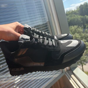 Svarta rockrunners - Svarta Valentino Garavani sneakers med coola silvriga detaljer och klassisk snörning. Snygg mix av skinn och textil, samt ikoniska nitar på hälen. Perfekta för dig som vill ha en lyxig och edgy look till vardags. Pris är ej hugget i sten, hör gärna av vid frågor!