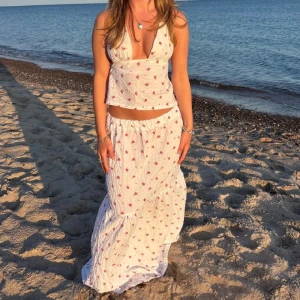 Vit långkjol med små rosa hjärtan - Supersöt vit långkjol med små rosa hjärtan, perfekt flowy och luftig för stranden eller varma dagar. Kjolen har resår i midjan och är gjord i ett lätt, skrynkligt bomullstyg som ger en avslappnad vibe.