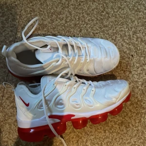 Nike Vapormax Plus vit/röd sneakers - Nike Vapormax Plus sneakers i vitt med röda detaljer och en riktigt fet röd genomskinlig sula. Snyggt mönstrad ovandel i syntetmaterial, klassisk snörning och ikonisk Nike-logga på sidan. Perfekta för dig som vill sticka ut med en modern och sportig look.