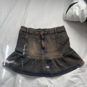 Svart jeanskjol med volangkant - Cool svart jeanskjol med volangkant längst ner och slitna detaljer. Kjolen har klassiska jeansfickor fram och bak, bälteshällor och knappstängning. Sitter snyggt på höften och har en lätt utsvängd form. Perfekt för dig som gillar en edgy vibe.