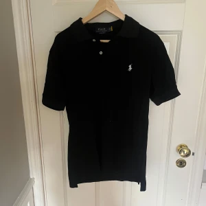 Svart pikétröja från Ralph Lauren - Klassisk svart pikétröja från Polo Ralph Lauren med vit broderad logga på bröstet. Tröjan har korta ärmar, krage och knäppning med två knappar. Tillverkad i bomull och har en slim passform som sitter snyggt på kroppen.