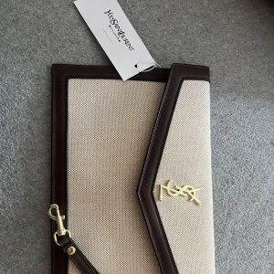YSL kuvertväska i beige och brunt - Stilren kuvertväska från YSL i beige canvas med mörkbruna skinnkanter och guldigt YSL-emblem. Väskan har en avtagbar handledsrem i brunt skinn och elegant asymmetrisk lockdesign. Perfekt för dig som vill ha en classy accessoar med lyxig känsla. Har inget äktehetsbevis, därav priset🥰