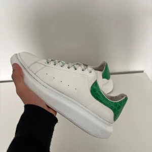 Alexander McQueen sneakers vit/grön - Snygga sneakers från Alexander McQueen i vitt skinn med grön, krokopräglad häl och matchande gröna detaljer på snörningen. Klassisk rund tå och platt sula ger en modern vibe. Perfekta för dig som vill sticka ut med stilrena men unika skor. Kvitto finns, extra snören