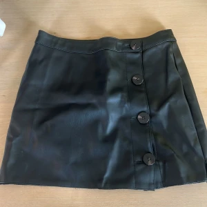 Svart skinnkjol med knappar - Snygg svart kortkjol i skinnimitation med stora knappar framtill. Kjolen har en clean look och passar perfekt till en cool streetstyle eller för att dressa upp outfiten. Enkel att matcha med både boots och sneakers.