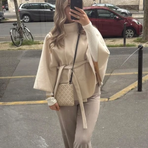 Beige poncho med bälte och guldknapp - Stilren beige poncho med hög krage, vida ärmar och knytbälte i midjan. Ponchon har en snygg guldknapp vid halsen och är tillverkad i ett mjukt material som ger en lyxig känsla. Perfekt för dig som gillar minimalistisk och trendig stil. Från Missäy