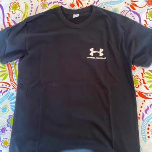 Svart t-shirt från Under Armour - Svart t-shirt från Under Armour med klassisk passform och rund hals. Diskret logga i vitt på bröstet ger en sportig vibe. Tillverkad i mjuk bomull som känns skön mot huden. Perfekt för dig som gillar enkel och stilren design.