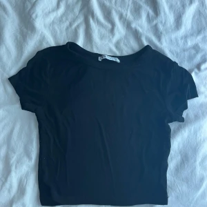 Svart croppad topp från Zara - Enkel och stilren svart croppad topp från Zara med rund halsringning och korta ärmar. Perfekt att matcha med jeans eller kjol för en clean look. Mjuk och stretchig känsla i materialet.