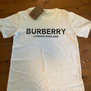 Vit Burberry t-shirt  - Vit t-shirt från Burberry med klassiskt svart logotyp-tryck 'BURBERRY LONDON ENGLAND' på bröstet. T-shirten har rund halsringning och korta ärmar. Tillverkad i mjuk bomull för en clean och stilren look. Helt ny!