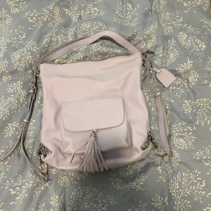 Ljuslila axelväska med tofs - Säljer en ljuslila axelväska i skinn med justerbar rem och snygg tofsdetalj framtill. Väskan har ett ytterfack med lock och dragkedja upptill, samt ett rymligt innerfack. Perfekt storlek för att få plats med allt du behöver till vardags.