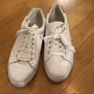 Vita sneakers från Gant med gulddetalj - Snygga vita sneakers från Gant med guldfärgade detaljer vid hälen och guldiga öljetter. Skorna har klassisk rund tå, breda vita snören och en diskret logga på sidan. Materialet är skinn och de har en platt sula. Endast andvända på studenten, därför lite skitiga