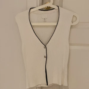 Vit ribbad linnetopp med svarta detaljer - Vit ribbad linnetopp från H&M med v-ringning och svarta kontrastsömmar längs kanten. Toppen är ärmlös och har två knappar framtill för en stilren look. Perfekt att styla med jeans eller kjol för en trendig outfit.