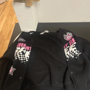 Svart bomberjacka med racingtryck - Svart bomberjacka med coolt racinginspirerat tryck i vitt och rosa framtill. Jackan har vita ränder längs ärmarna, tryckknappar och ribbade muddar. Materialet känns mjukt och bekvämt, perfekt för dig som gillar streetstyle och sportiga vibes.