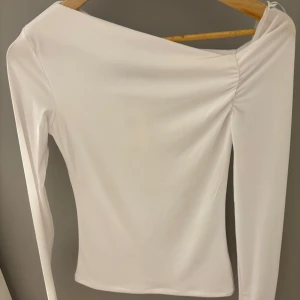 Vit topp från BikBok - Säljer en vit långärmad topp från BikBok i omlottmodell med lätt rynk vid axeln. Toppen har en stilren, asymmetrisk offshoulder-skärning och är gjord i ett mjukt, stretchigt material som sitter snyggt på kroppen. Nypris 300kr, prislapp på