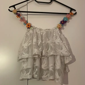 Vit volangig blus med spets och blommiga band - Supersöt vit blus med lager av volanger och spetsdetaljer. Axelbanden är dekorerade med färgglada blommor på ena toppen och vita blommor på den andra, vilket ger en unik och lekfull look. Perfekt för sommarens alla tillfällen när du vill sticka ut lite extra. Det blir då 2 toppar