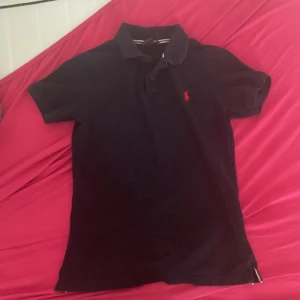 Svart pikétröja från Ralph Lauren - Klassisk svart pikétröja från Ralph Lauren med röd broderad logga på bröstet. Tröjan har korta ärmar, krage och knappar framtill. Tillverkad i bomull för en skön och stilren känsla. Perfekt för dig som gillar en tidlös och sportig look.