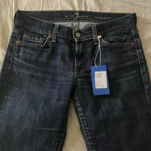 Mörkblå bootcut jeans 7 For All Mankind - Snygga mörkblå jeans från 7 For All Mankind, modell Roxanne. Klassisk femficksdesign, raka ben med bootcut och slits nertill. Jeansen har en mörk tvätt och subtila slitningar. Perfekta för dig som gillar en tidlös och stilren look. Kan tilllägga att dom är mer som skinny jeans men för någon med smalare vader sitter dom bottcut 