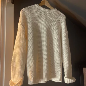 Vit chunky stickad tröja - Säljer en vit chunky stickad tröja med grov struktur och ribbade muddar vid ärmslut och nederkant. Tröjan har en avslappnad passform och långa ärmar, perfekt för lager-på-lager under kyliga dagar. Finns en tråd som gått upp men går säkert bra att bara klippa av 